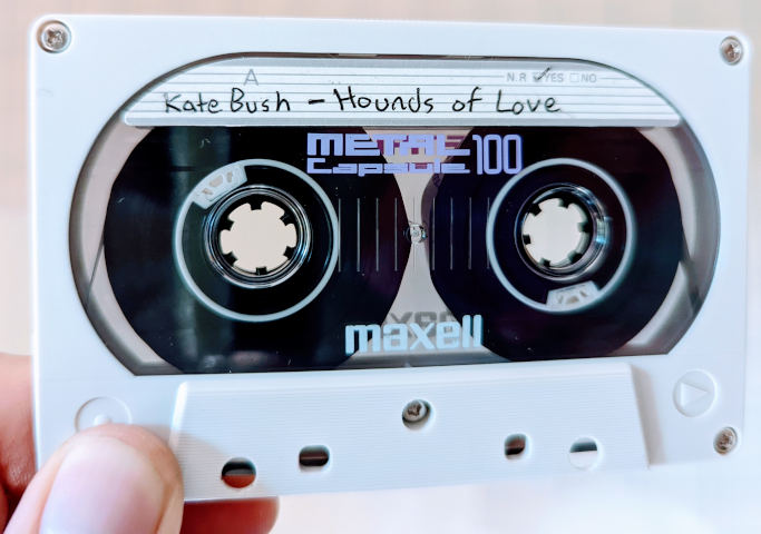 A Maxel Metal Capsule C-100 cassette. The shell is off-white with rounded corners and a very large oval window. The label indicates that Hounds of Love by Kate Bush is recorded to Side A